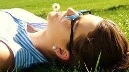 Eine Frau liegt mit Sonnenbrille im Gras, im Mund ein Gänseblümchen