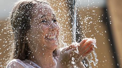 Eine Frau steht nass in einem Wasserstrahl, der kommt aus einem Brunnen